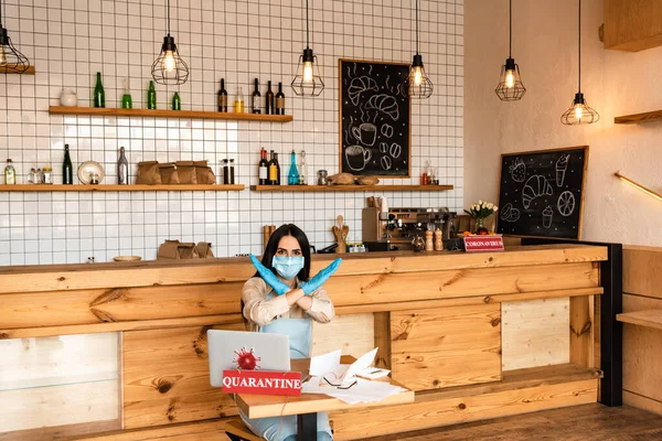 Cafe owner in medical mask showing no sign at table with laptop, papers and card with quarantine inscription — Stock Photo