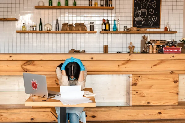 Propietario del café en guantes de látex con las manos cerradas sobre la cabeza cerca de documentos, calculadora, gafas y portátil en la mesa - foto de stock