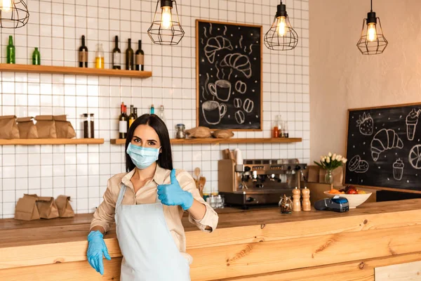 Propietario de café en máscara médica y guantes de látex mirando a la cámara y mostrando como signo cerca de la mesa - foto de stock