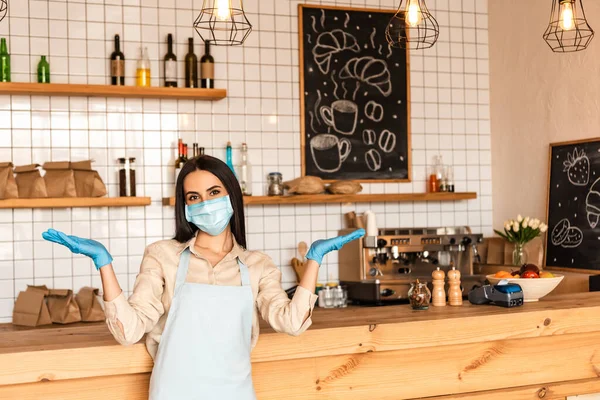 Cafébesitzer mit offenen Armen in medizinischer Maske und Latexhandschuhen blickt in die Kamera am Tisch — Stockfoto