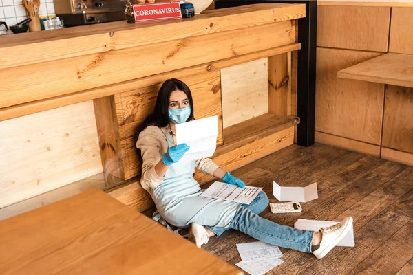 Vista ad alto angolo del proprietario del caffè scioccato in maschera medica guardando carta vicino calcolatrice e tavolo sul pavimento — Foto stock