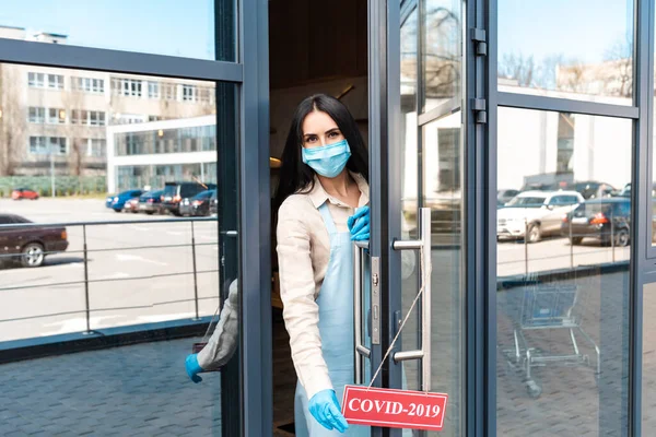 Proprietário do café em máscara médica perto da porta olhando para a câmera e cartão de toque com letras covid-2019 na rua — Fotografia de Stock