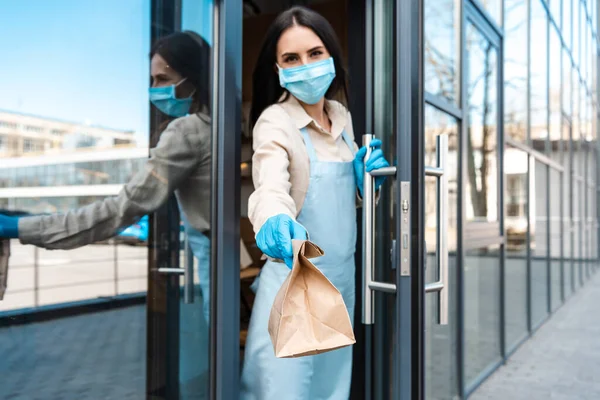 Dono do café em máscara médica tocando maçaneta da porta, olhando para a câmera e mostrando saco de papel na rua — Fotografia de Stock