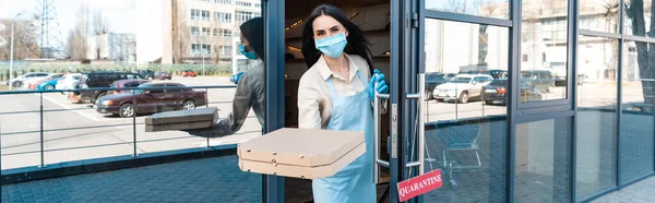 Imagen horizontal del dueño de la cafetería en máscara médica cerca de la puerta con tarjeta con letras de cuarentena que muestran las cajas y mirando a la cámara en la calle - foto de stock