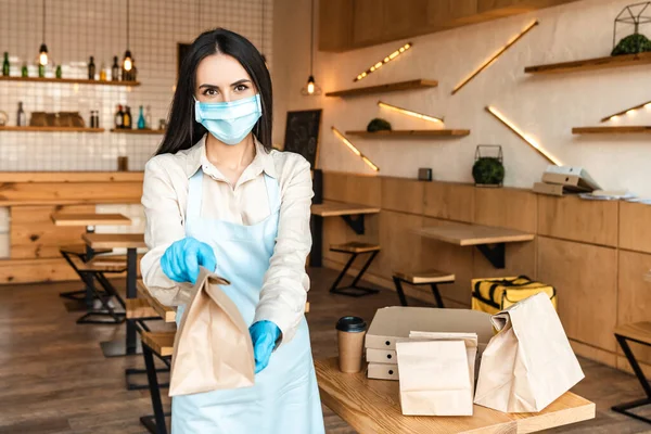 Cafe owner in medical mask showing paper bag and looking at camera near table with disposable cup of coffee and boxes — Stock Photo