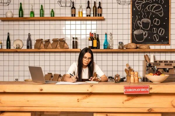 Concentrated cafe owner with pen and papers using calculator at table with laptop and card with coronavirus inscription — Stock Photo