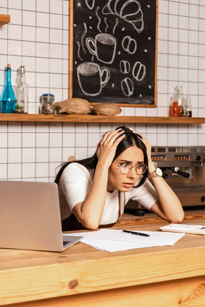 Schockierter Café-Besitzer schaut mit Stift und Laptop auf Taschenrechner neben Zetteln am Tisch — Stockfoto