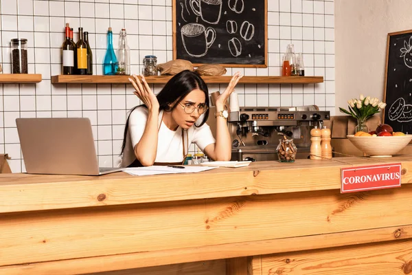 Propriétaire de café choqué regardant calculatrice près de papiers et ordinateur portable à table avec carte avec inscription coronavirus — Photo de stock