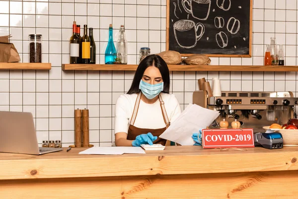 Cafe owner in medical mask with papers using calculator near laptop and card with covid-2019 lettering on table — Stock Photo