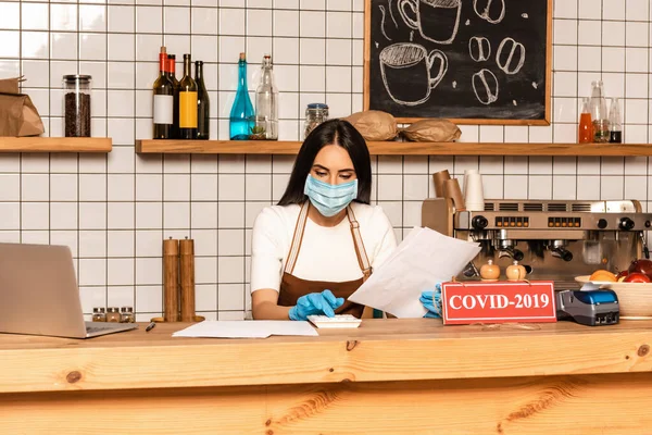 Propietario del café en máscara médica con papeles usando la calculadora cerca del ordenador portátil y la tarjeta con letras covid-2019 en la mesa - foto de stock