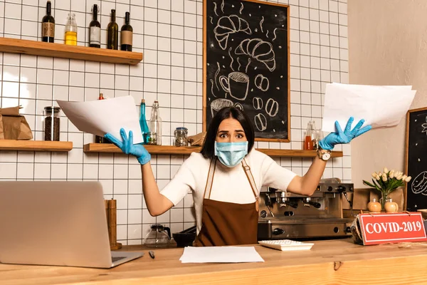 Worried cafe owner in medical mask with papers looking at camera near table with laptop, calculator and card with covid-2019 inscription — Stock Photo