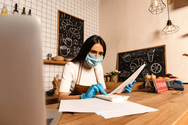 Worried cafe owner in medical mask with papers using calculator near laptop at table — Stock Photo