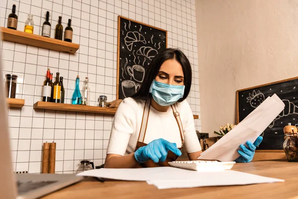 Concentrated cafe owner in medical mask with papers using calculator near laptop at table — Stock Photo