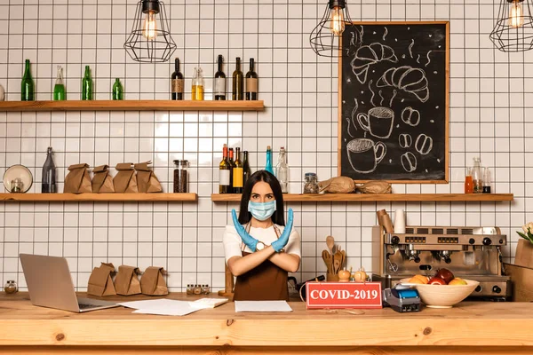 Cafe owner in medical mask showing no sign near table with papers, card with covid-2019 lettering, payment terminal and bowl with fruits — Stock Photo