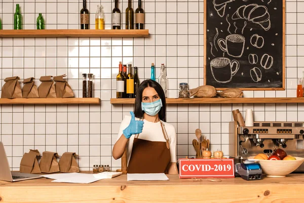 Cafe owner in medical mask showing like sign near table with papers, card with covid-2019 inscription, payment terminal and bowl with fruits — Stock Photo