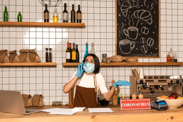 Cafe owner in medical mask talking on smartphone near table with laptop, papers, payment terminal and bowl with fruits — Stock Photo