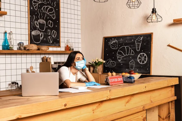 Thoughtful cafe owner near table with laptop, papers, card with covid-2019 lettering, payment terminal and bowl with fruits — Stock Photo