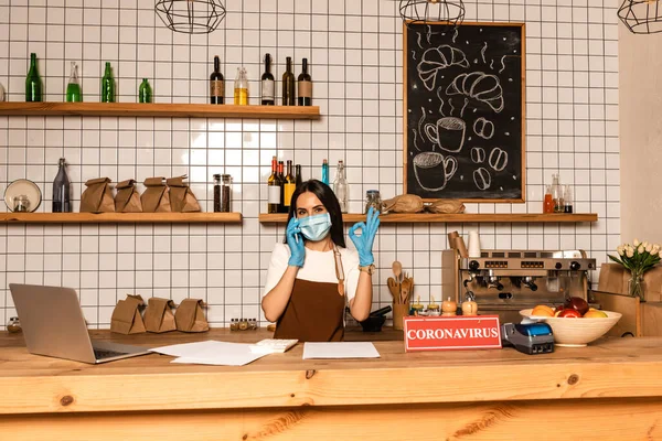 Propietario del café hablando en el teléfono inteligente y mostrando signo bien cerca de la mesa con el ordenador portátil, papeles, tarjeta con letras coronavirus, terminal de pago y tazón con frutas - foto de stock