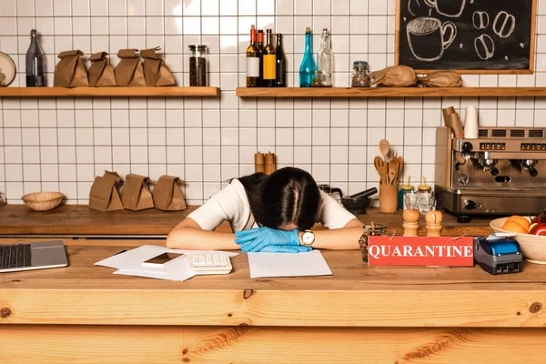 Propietario del café con la cabeza en las manos en la mesa con gadgets, papeles, tarjeta con letras de cuarentena, terminal de pago y tazón con frutas - foto de stock