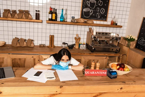 Vista de alto ángulo del dueño de la cafetería en máscara médica en la mesa con teléfono inteligente, papeles, calculadora, tarjeta con letras de cuarentena, terminal de pago y frutas - foto de stock