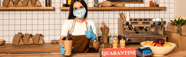 Horizontal image of cafe owner in medical mask showing like sign and paper cup of coffee near table with card with quarantine inscription — Stock Photo