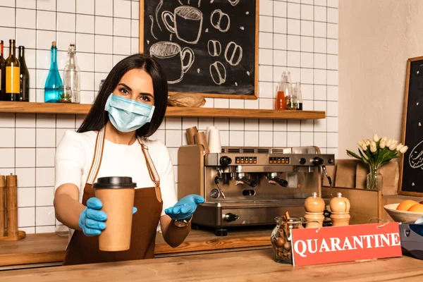 Cafe owner in medical mask pointing at paper cup of coffee near table with salt and pepper mills, jar and card with quarantine inscription — Stock Photo