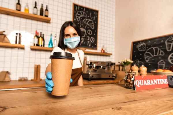 Selective focus of cafe owner in medical mask showing paper cup of coffee near table with card with quarantine inscription — Stock Photo