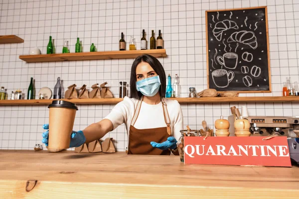 Cafe owner in medical mask looking at camera and pointing at paper cup of coffee near table with card with quarantine inscription — Stock Photo
