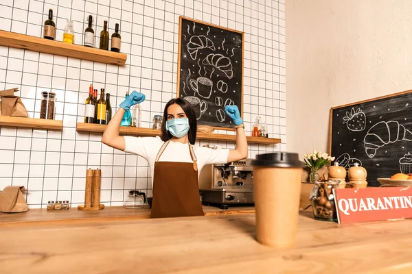 Selective focus of disposable cup of coffee near card with quarantine lettering on table and cafe owner in medical mask with hands in air — Stock Photo