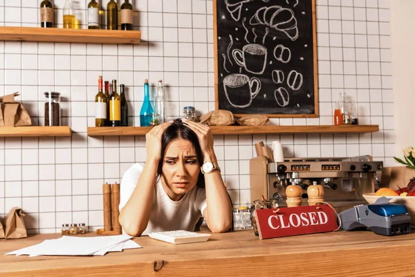 Worried cafe owner at table with calculator, documents, card with closed lettering and payment terminal — Stock Photo