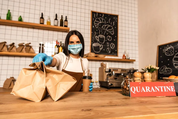 Cafe owner in medical mask with disposable cup of coffee showing paper bags near table with card with closed inscription — Stock Photo