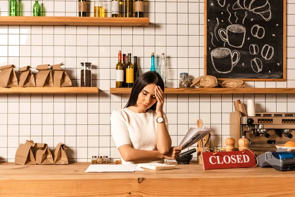 Stressed cafe owner with closed eyes holding papers near table with calculator, card with closed lettering and payment terminal — Stock Photo