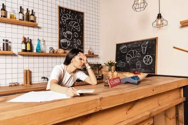 Cafébesitzer hält Taschenrechner in der Nähe von Papieren, Karte mit geschlossenem Schriftzug und Zahlungsterminal auf Tisch — Stockfoto