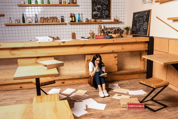 Propriétaire de café choqué regardant calculatrice et assis près de la table, papiers et carte avec lettrage coronavirus sur le sol — Photo de stock