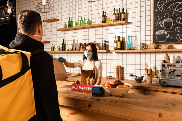 Cafe owner in medical mask giving paper bag to courier near table with payment terminal, bowl with fruits and card with quarantine inscription — Stock Photo