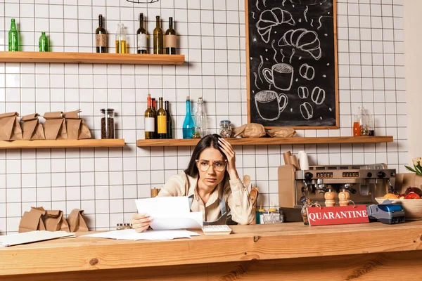 Stressed cafe owner touching head and looking at papers near card with quarantine lettering and payment terminal near table — Stock Photo