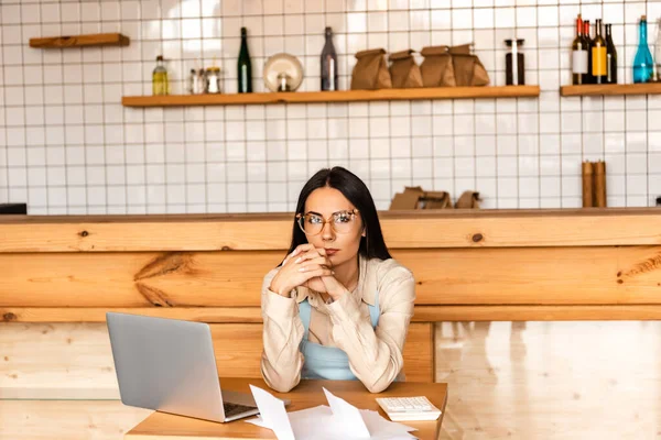 Thoughtful cafe owner with clenched hands near laptop, papers and calculator at table — Stock Photo