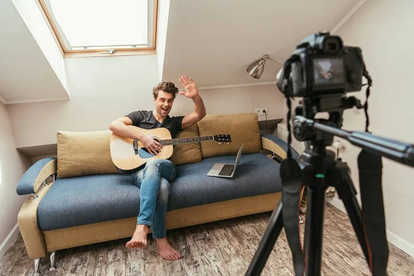 Foyer sélectif du vlogger souriant tenant la guitare et agitant la main à l'appareil photo numérique sur le trépied — Photo de stock