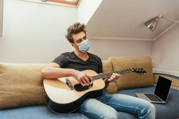 Young man in protective mask playing guitar while sitting on sofa near laptop with blank screen — Stock Photo