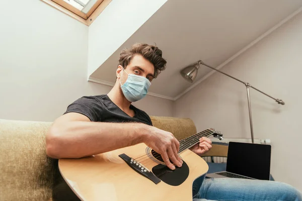 Visão de baixo ângulo do jovem na máscara médica tocando guitarra e olhando para a câmera enquanto sentado no sofá perto do laptop com tela em branco — Fotografia de Stock