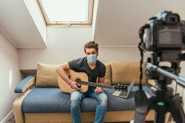 Selective focus of young vlogger in medical mask playing guitar while looking at digital camera on tripod — Stock Photo