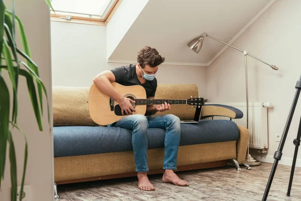 Молодий чоловік в медичній масці грає на гітарі, сидячи на дивані біля ноутбука — стокове фото