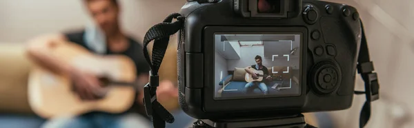Selective focus of young vlogger playing guitar while looking at digital camera, horizontal image — Stock Photo