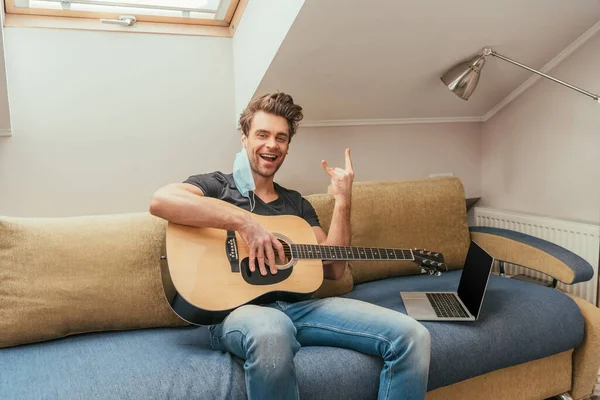 Uomo allegro con maschera medica sull'orecchio che tiene la chitarra e mostra il segno della roccia mentre si siede sul divano vicino al computer portatile con schermo bianco — Foto stock