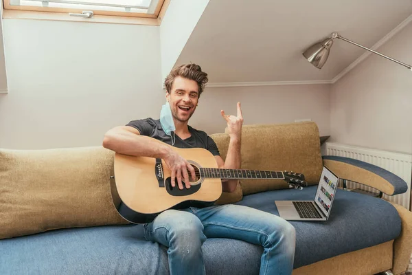 KIEW, UKRAINE - 13. APRIL 2019: fröhlicher Mann mit Schutzmaske am Ohr, der eine Gitarre hält und Rockzeichen zeigt, während er auf dem Sofa neben dem Laptop sitzt und Youtube auf dem Bildschirm zeigt — Stockfoto