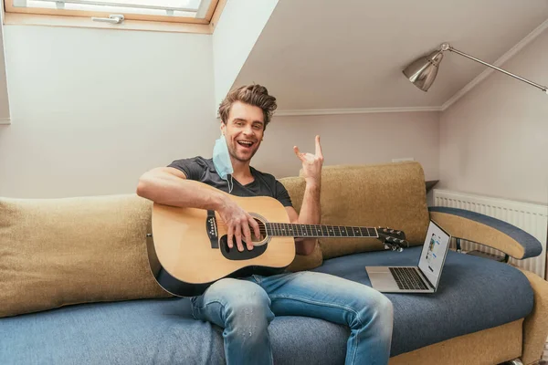 KYIV, UCRANIA - 13 de ABRIL de 2019: hombre alegre con máscara médica en la oreja sosteniendo la guitarra y mostrando el cartel de rock mientras está sentado en el sofá cerca de la computadora portátil con Instagram en la pantalla - foto de stock