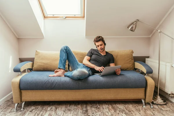 Красивый мужчина в гарнитуре лежит на диване рядом с ноутбуком, работая дома в мансарде — стоковое фото
