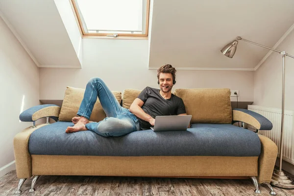 Fröhlicher Mann mit Headset auf Sofa neben Laptop liegend und in Kamera blickend — Stockfoto