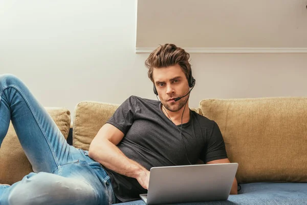 Hombre serio en auriculares tumbado en el sofá cerca de la computadora portátil y mirando a la cámara - foto de stock