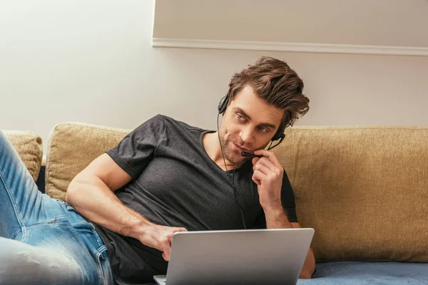 Hombre escéptico en auriculares tumbado en el sofá cerca de la computadora portátil, mientras que el trabajo en casa - foto de stock